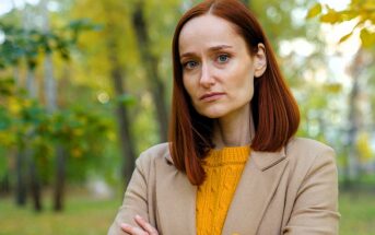 Woman with red hair wearing a beige coat and yellow sweater stands outdoors with arms crossed. She appears thoughtful, surrounded by blurred green and yellow foliage in a park setting.