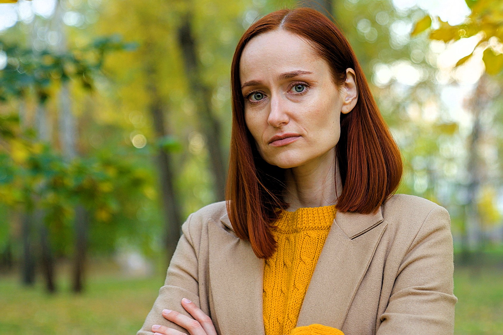 Woman with red hair wearing a beige coat and yellow sweater stands outdoors with arms crossed. She appears thoughtful, surrounded by blurred green and yellow foliage in a park setting.