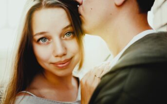 A woman with long hair smiles softly while a man kisses her forehead. She rests her hand on his chest, displaying a ring. The background is softly lit, creating a warm and intimate atmosphere.