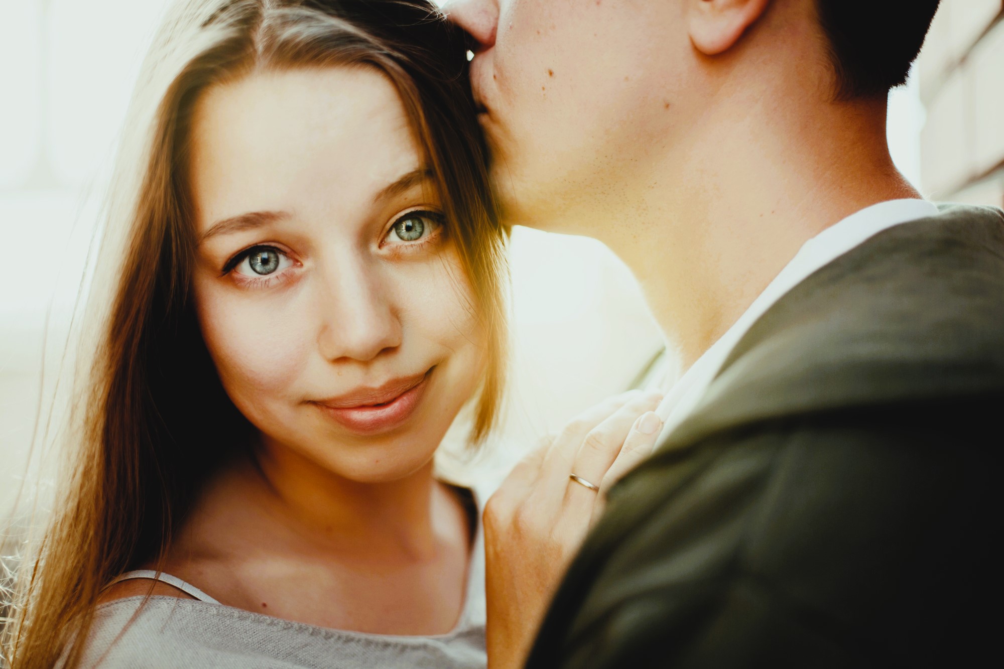 A woman with long hair smiles softly while a man kisses her forehead. She rests her hand on his chest, displaying a ring. The background is softly lit, creating a warm and intimate atmosphere.