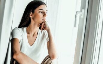 A woman with long dark hair sits by a window, resting her chin on her hand. She is wearing a white T-shirt and appears to be deep in thought, gazing outside. The lighting is soft, adding a contemplative mood to the scene.