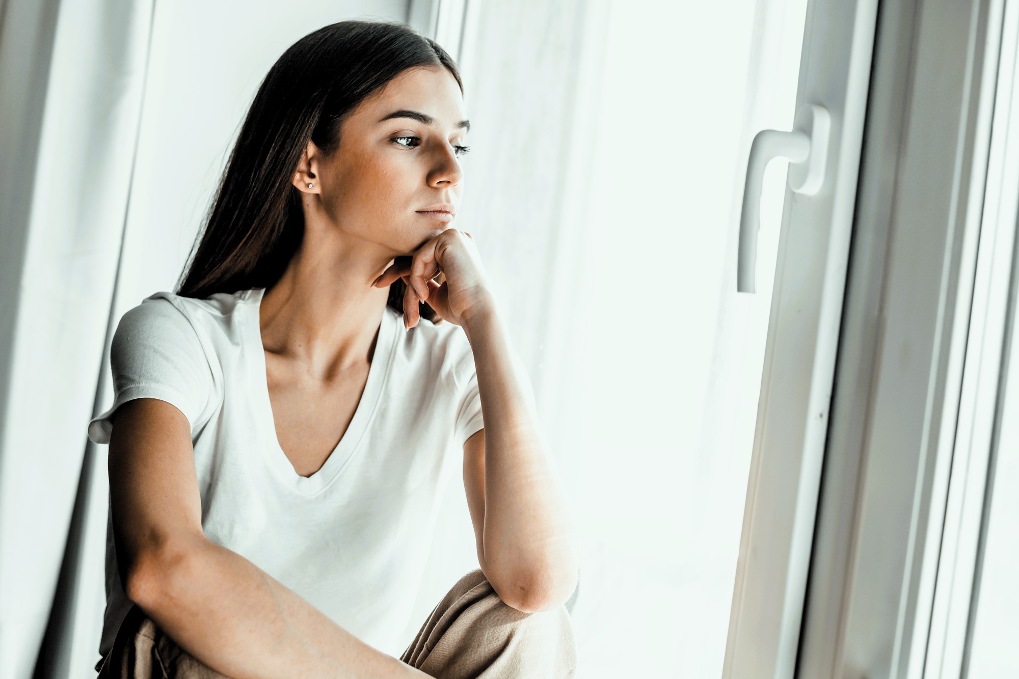 A woman with long dark hair sits by a window, resting her chin on her hand. She is wearing a white T-shirt and appears to be deep in thought, gazing outside. The lighting is soft, adding a contemplative mood to the scene.