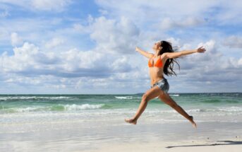 A woman in an orange bikini top and denim shorts joyfully leaps on a sandy beach. The ocean waves gently roll in the background under a partly cloudy sky.