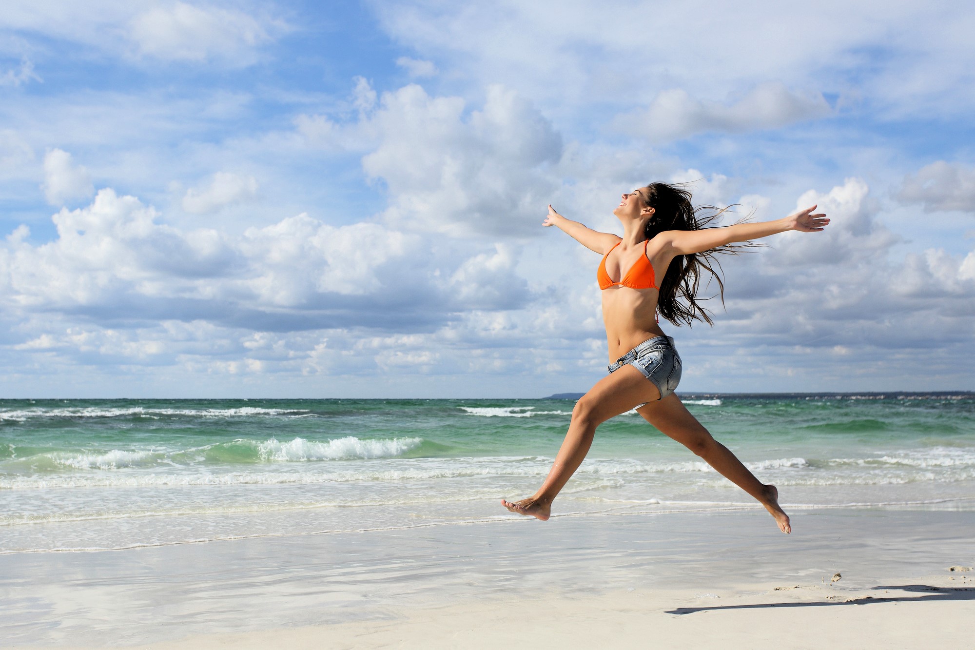 A woman in an orange bikini top and denim shorts joyfully leaps on a sandy beach. The ocean waves gently roll in the background under a partly cloudy sky.