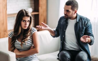 A young woman with long hair, wearing a striped shirt, sits on a couch with arms crossed, looking upset. A man in a denim shirt and white t-shirt sits beside her, gesturing with his hands, appearing to be in a serious conversation.