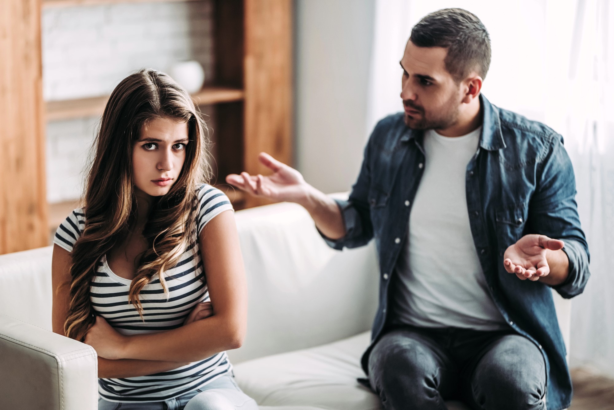 A young woman with long hair, wearing a striped shirt, sits on a couch with arms crossed, looking upset. A man in a denim shirt and white t-shirt sits beside her, gesturing with his hands, appearing to be in a serious conversation.