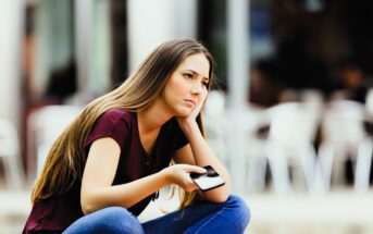 A woman with long brown hair, in a maroon shirt and blue jeans, sits outdoors with a smartphone in hand. She appears thoughtful or concerned, resting her face on her other hand. There are blurred chairs and tables in the background.