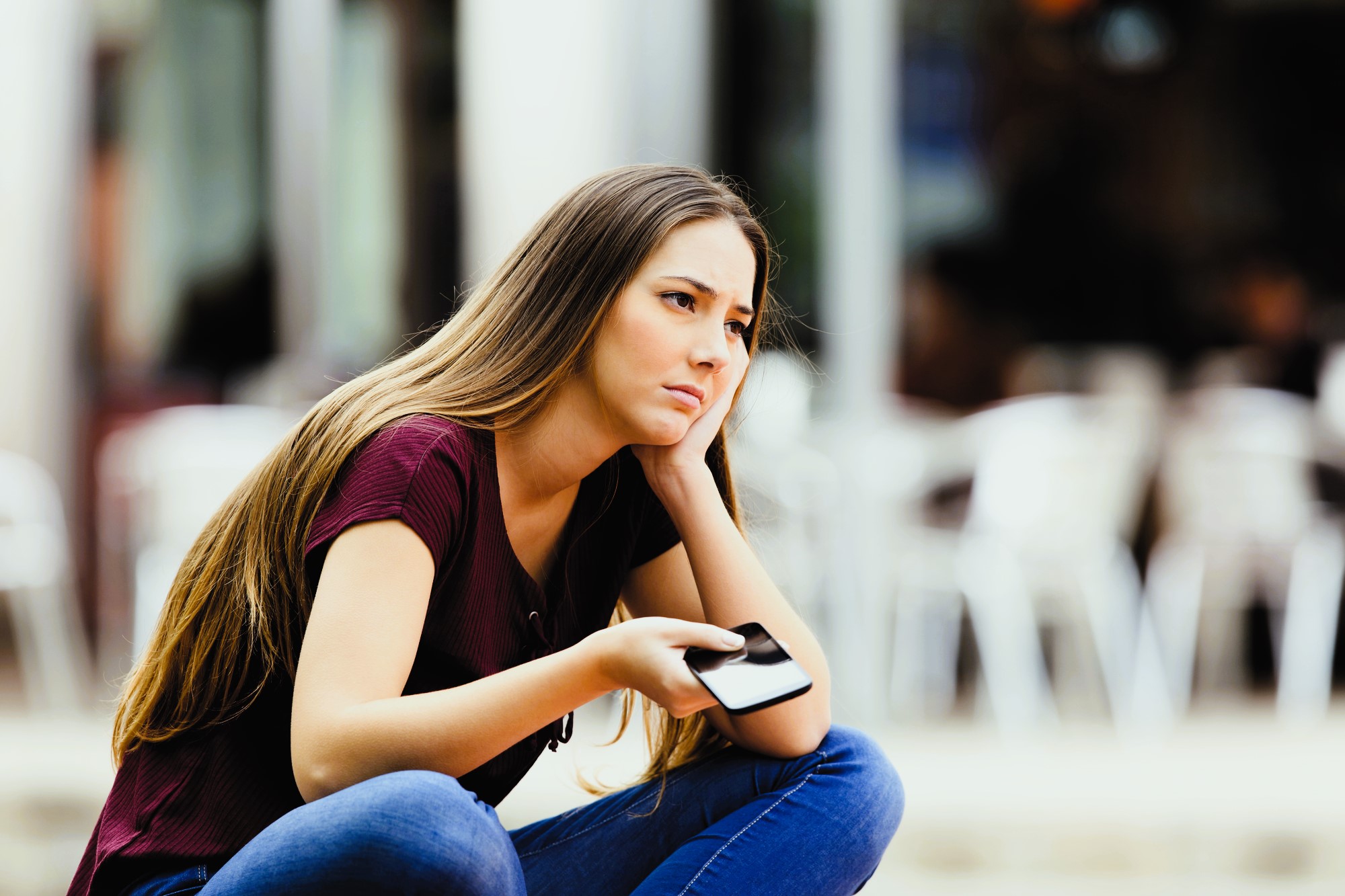 A woman with long brown hair, in a maroon shirt and blue jeans, sits outdoors with a smartphone in hand. She appears thoughtful or concerned, resting her face on her other hand. There are blurred chairs and tables in the background.