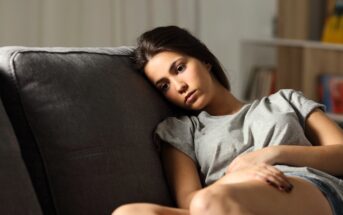 Young woman in a gray t-shirt sits on a couch, looking pensive and thoughtful. She leans her head against the backrest, with soft lighting and a blurred background of shelves and a light wall.