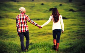 A couple is walking hand in hand through a lush green field. The man wears a checkered shirt and beanie, and the woman is in a white sweater and boots. Both are facing away, enjoying the serene landscape.