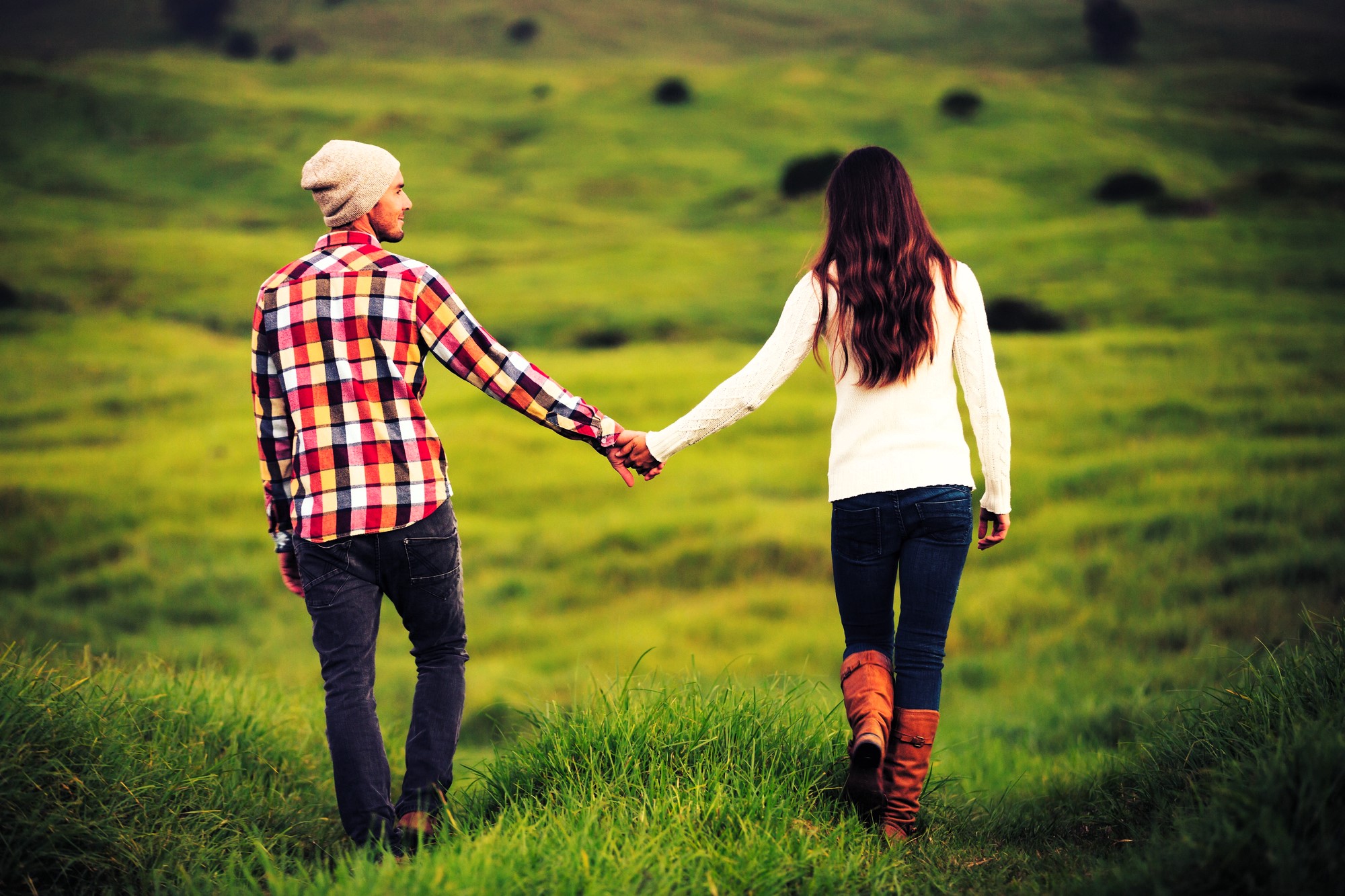 A couple is walking hand in hand through a lush green field. The man wears a checkered shirt and beanie, and the woman is in a white sweater and boots. Both are facing away, enjoying the serene landscape.