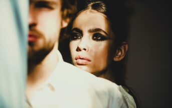 A woman with dramatic eye makeup and intense gaze stands partly obscured behind a man in a white shirt, bathed in warm sunlight. Only the side of the man's face is visible, creating a shadowy and mysterious atmosphere.