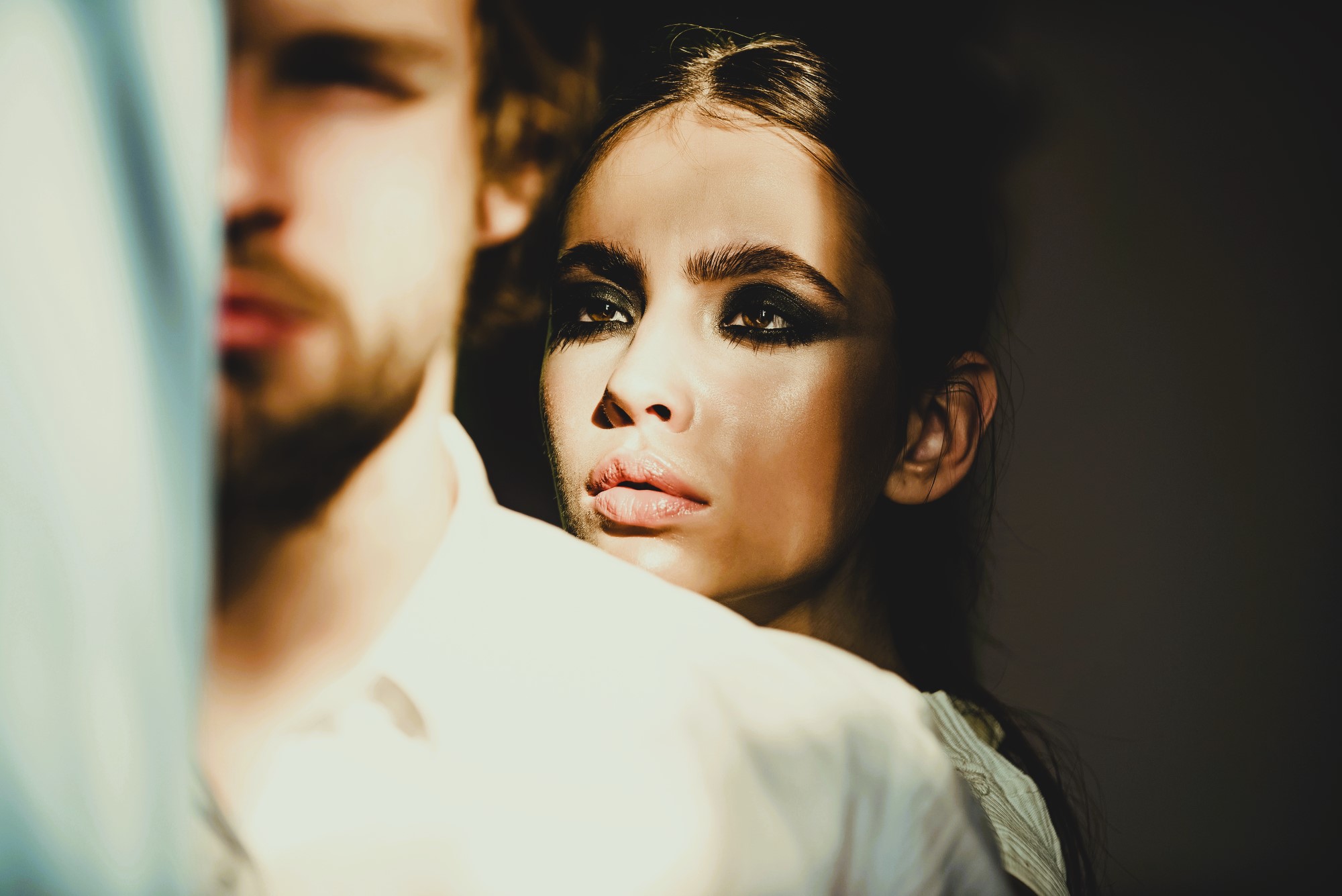 A woman with dramatic eye makeup and intense gaze stands partly obscured behind a man in a white shirt, bathed in warm sunlight. Only the side of the man's face is visible, creating a shadowy and mysterious atmosphere.