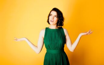 A woman in a green dress stands against a bright yellow background, with a puzzled expression and her arms raised in a questioning gesture.