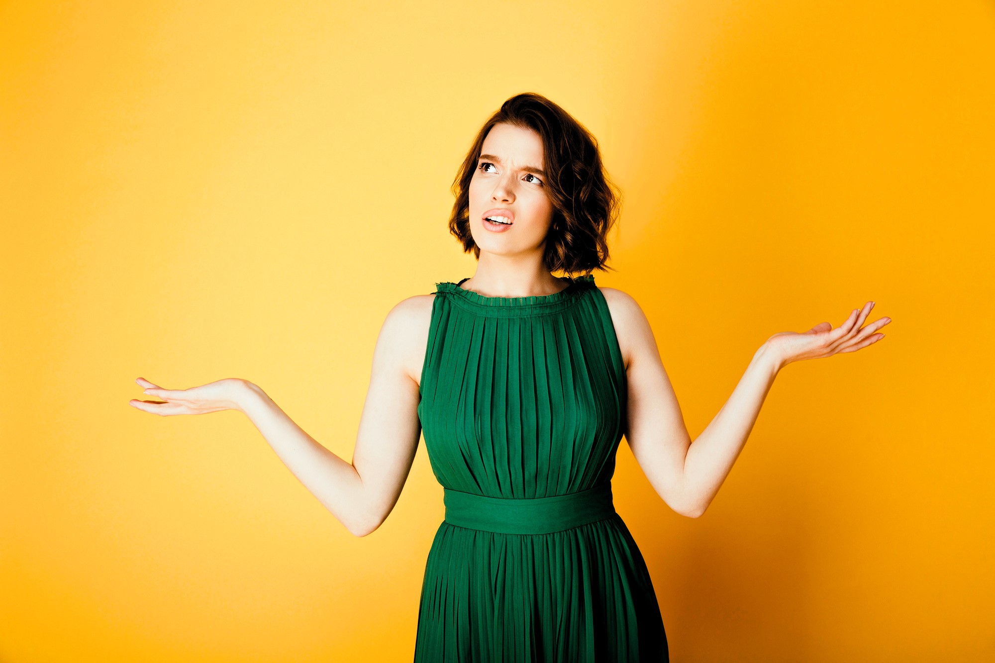 A woman in a green dress stands against a bright yellow background, with a puzzled expression and her arms raised in a questioning gesture.