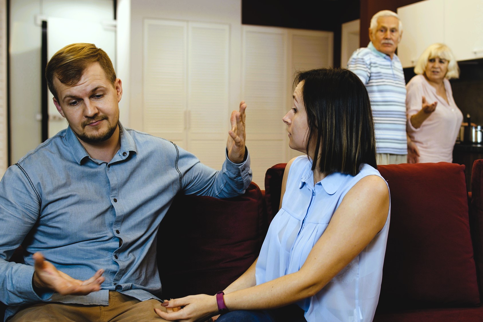 A young couple sits on a couch, engaged in a heated discussion, their hands gesturing expressively. In the background, an older couple stands in the kitchen, observing with concerned expressions. The setting is a modern living room.