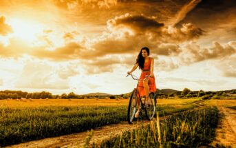 A woman in a red dress rides a bicycle on a dirt path through a grassy field under a dramatic, golden sky with clouds and sun. Hills and trees are visible in the background.