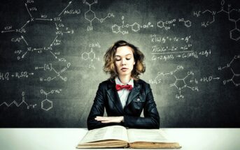 A person with curly hair and a red bow tie sits at a desk with an open book. They are wearing a black suit and appear focused. Behind them, a blackboard displays various chemical formulas and equations.