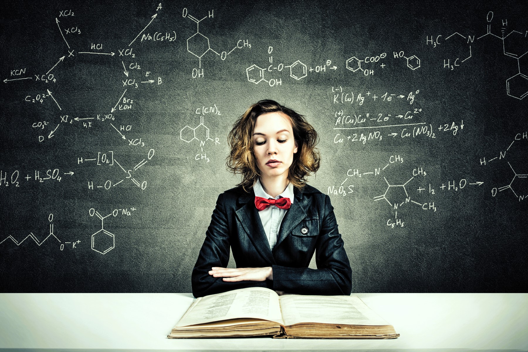 A person with curly hair and a red bow tie sits at a desk with an open book. They are wearing a black suit and appear focused. Behind them, a blackboard displays various chemical formulas and equations.