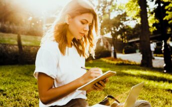 A person with long hair sits on grass in a park, writing in a notebook. They wear a white t-shirt and are next to an open laptop. Sunlight filters through trees, creating a bright, serene atmosphere.