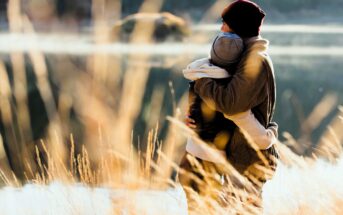 A couple stands hugging near a serene lake, surrounded by tall grasses. They wear warm jackets and beanies, looking out across the water on a bright, chilly day.