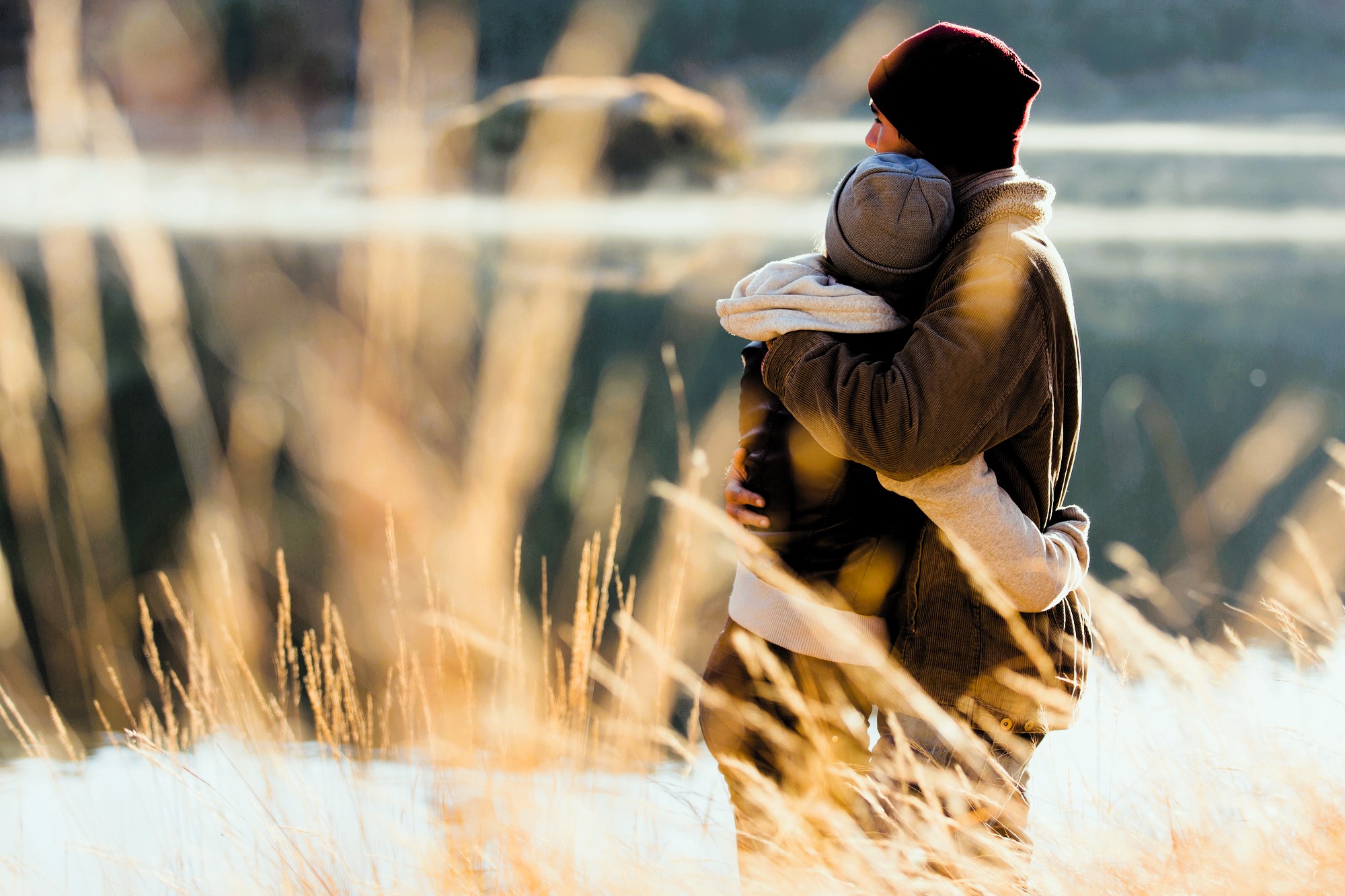 A couple stands hugging near a serene lake, surrounded by tall grasses. They wear warm jackets and beanies, looking out across the water on a bright, chilly day.