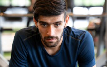 A man with a beard is wearing a dark blue shirt, looking intently at the camera. He appears to be in a gym, with exercise equipment in the blurred background.