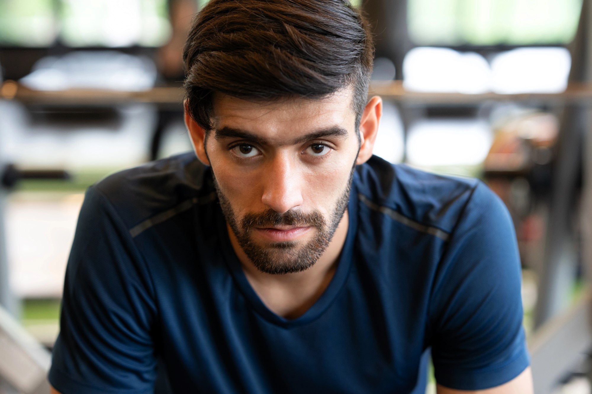 A man with a beard is wearing a dark blue shirt, looking intently at the camera. He appears to be in a gym, with exercise equipment in the blurred background.