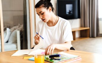 A woman with dark hair in a bun is sitting at a wooden table, writing in an open notebook with a blue pen. She has several colorful books and a glass of orange juice in front of her. A TV is visible in the background.