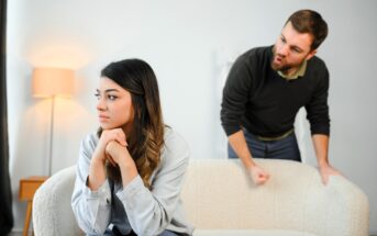 A woman sits on a couch, looking away with a thoughtful expression, while a man angrily gestures behind her, leaning over the couch. The room has a lamp in the background.