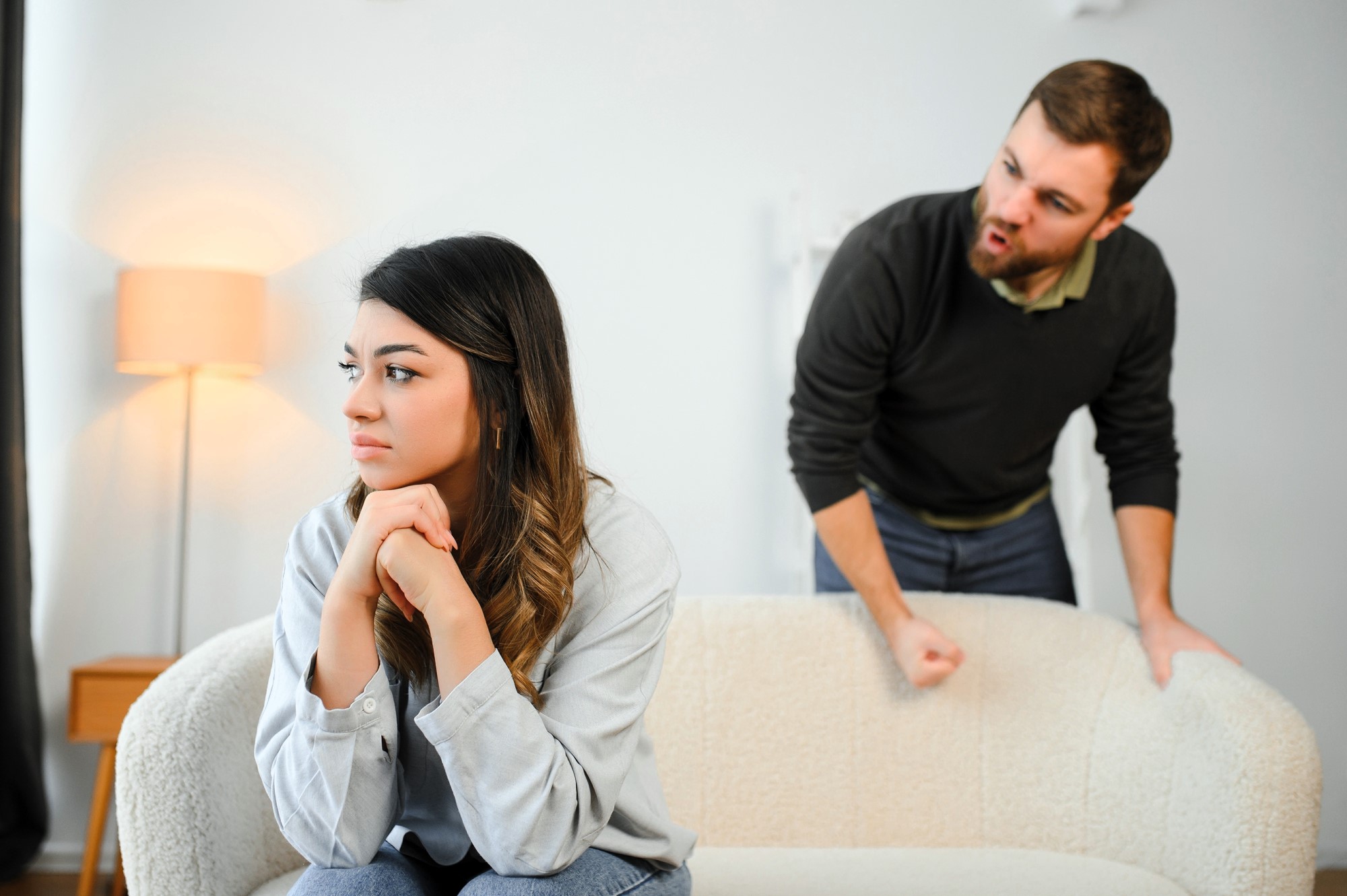 A woman sits on a couch, looking away with a thoughtful expression, while a man angrily gestures behind her, leaning over the couch. The room has a lamp in the background.