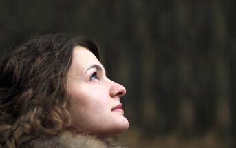 A woman with curly hair and a fur-lined coat looks upward against a blurred, dark green forest background. Her face is in profile, and her expression is contemplative.