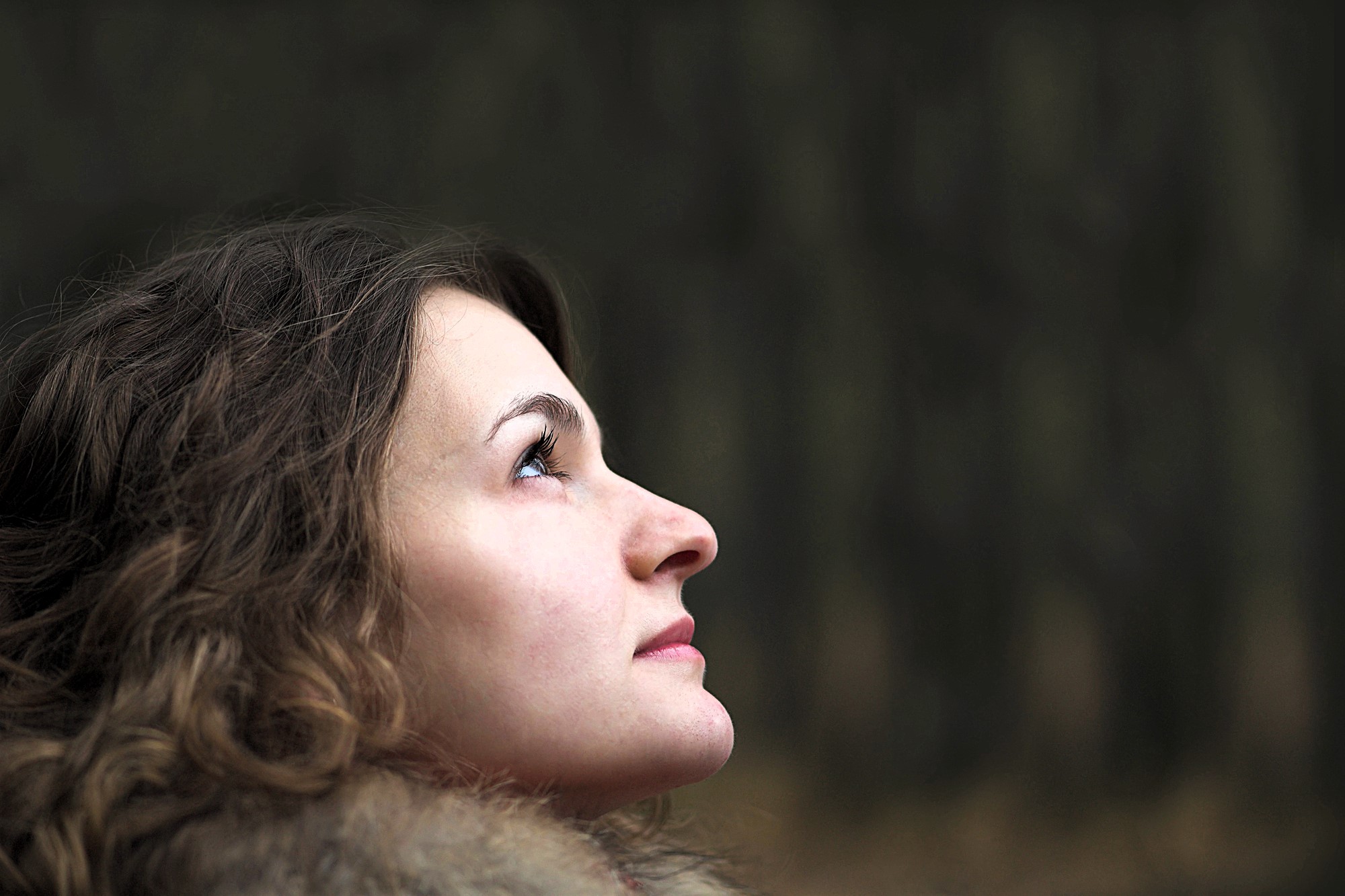 A woman with curly hair and a fur-lined coat looks upward against a blurred, dark green forest background. Her face is in profile, and her expression is contemplative.