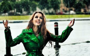 A woman in a bright green raincoat smiles joyfully while standing in the rain. She has long, wet hair and her arms are raised. The background shows a blurred outdoor setting with trees and pavement.