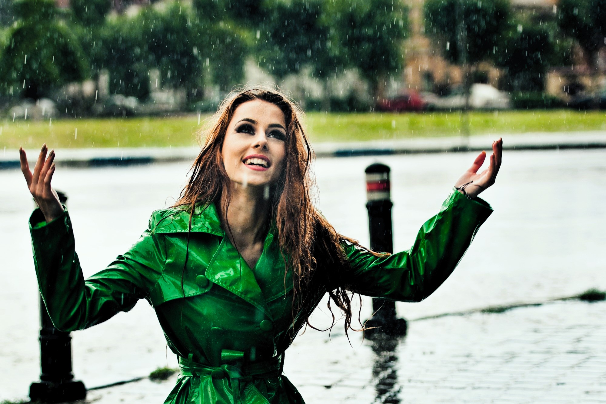 A woman in a bright green raincoat smiles joyfully while standing in the rain. She has long, wet hair and her arms are raised. The background shows a blurred outdoor setting with trees and pavement.