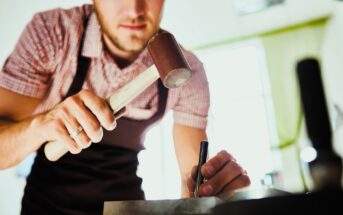 A person wearing an apron is using a mallet and a metal tool for crafting. They are focusing intently on their work, which is taking place on a table in a well-lit room.