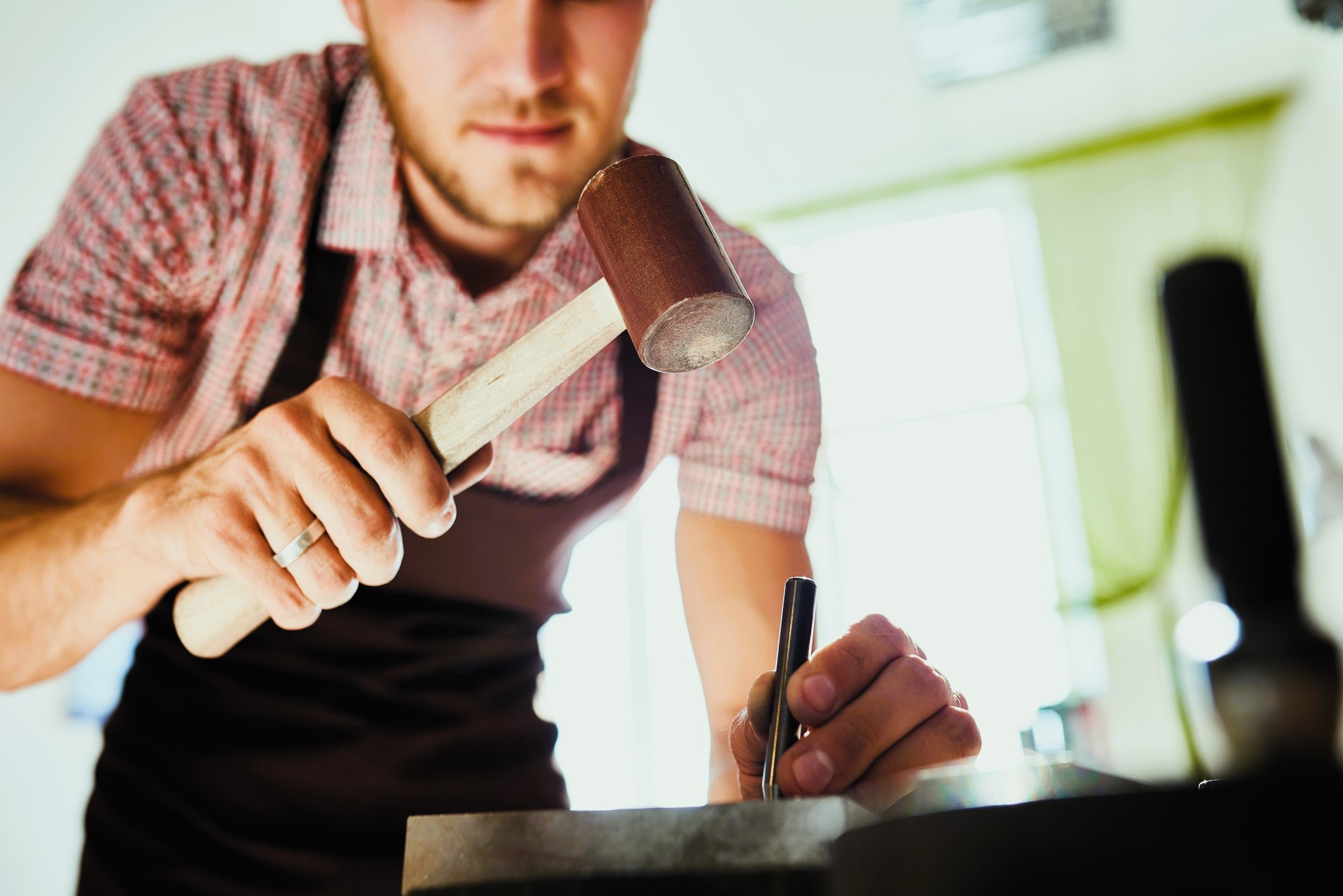 A person wearing an apron is using a mallet and a metal tool for crafting. They are focusing intently on their work, which is taking place on a table in a well-lit room.