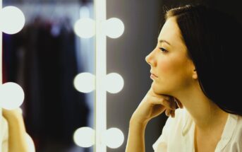 A woman with long hair is gazing at her reflection in a mirror surrounded by bright round lights. She rests her chin on her hand and is wearing a white top. In the background, blurred clothing can be seen.