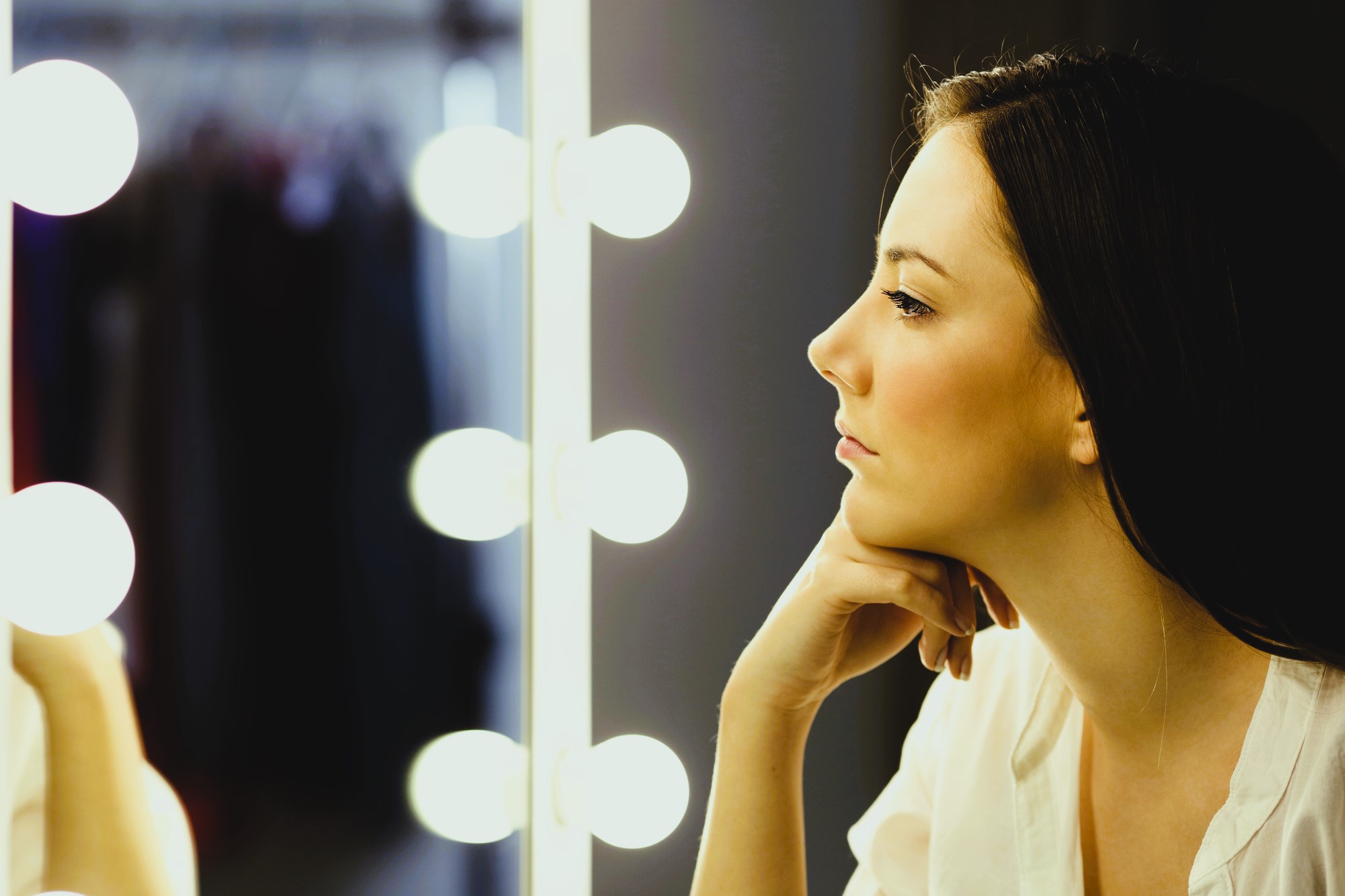 A woman with long hair is gazing at her reflection in a mirror surrounded by bright round lights. She rests her chin on her hand and is wearing a white top. In the background, blurred clothing can be seen.