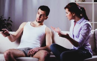 A woman in a light blue shirt is sitting on a couch, gesturing expressively towards a man in a white tank top and shorts who is holding a remote control. The man looks away, appearing uninterested.