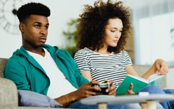 A man and woman sit on a couch. The man is holding a game controller and looking at a TV screen, while the woman sits beside him reading a book. The setting is a cozy living room with soft lighting.