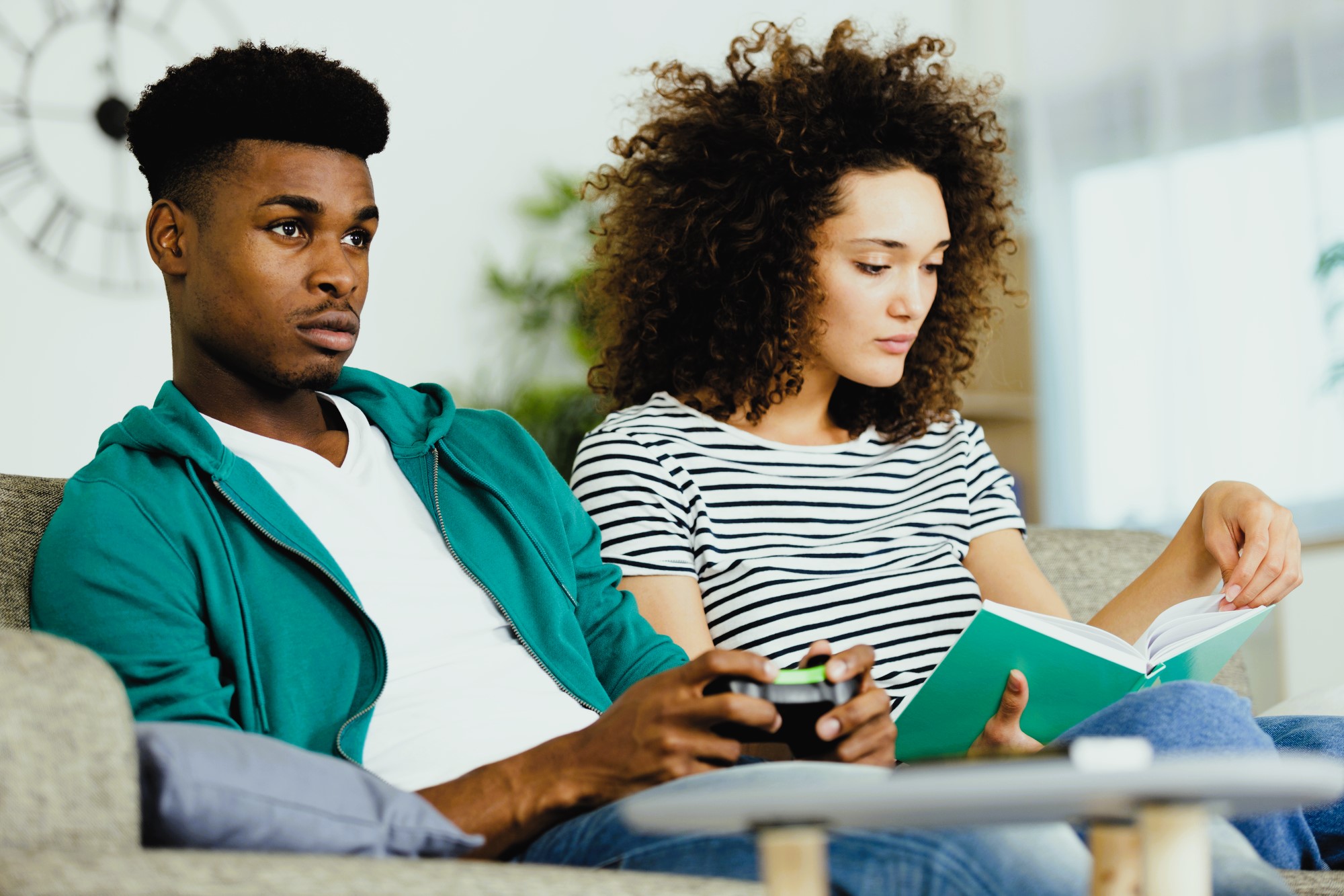 A man and woman sit on a couch. The man is holding a game controller and looking at a TV screen, while the woman sits beside him reading a book. The setting is a cozy living room with soft lighting.