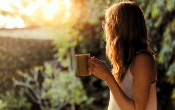 A person with long hair holding a coffee mug stands outdoors, gazing at a bright, sunlit scene surrounded by greenery. The soft sunlight creates a warm, golden glow.