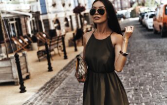 A woman in a sleeveless olive green dress and sunglasses is walking on a cobblestone street. She holds a patterned clutch and wears a watch. A row of buildings and parked vehicles line the street in the background.