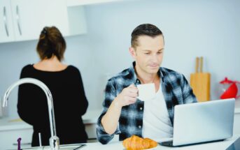 A man sits at a kitchen counter with a laptop and a croissant, holding a cup. He wears a plaid shirt. A woman in a black top stands with her back turned, near the kitchen sink. The setting is a modern, bright kitchen.