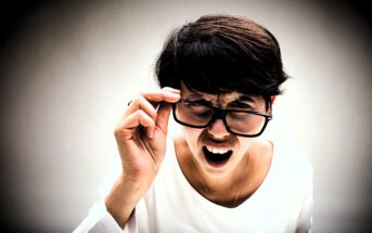 A person with short dark hair and a white shirt is adjusting their glasses while leaning forward and squinting, with a perplexed or expressive face against a plain background.