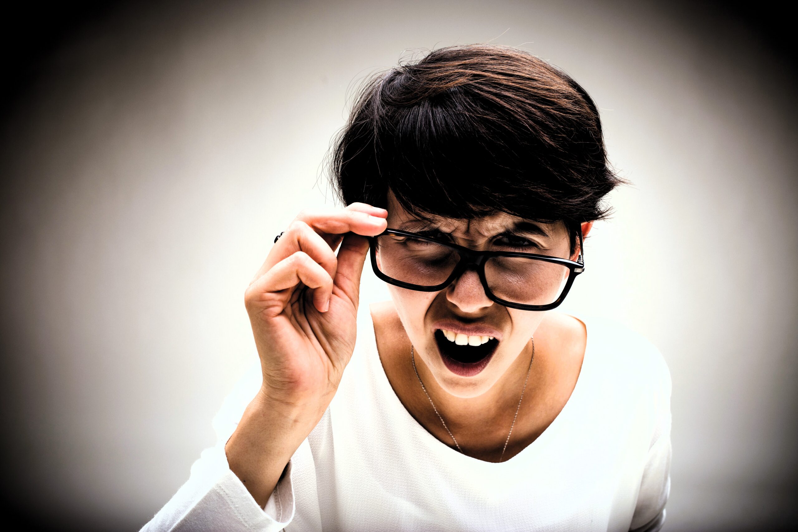 A person with short dark hair and a white shirt is adjusting their glasses while leaning forward and squinting, with a perplexed or expressive face against a plain background.