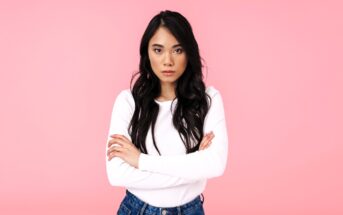 A woman with long dark hair stands against a pink background, wearing a white sweater and blue jeans. She has her arms crossed and a serious expression.