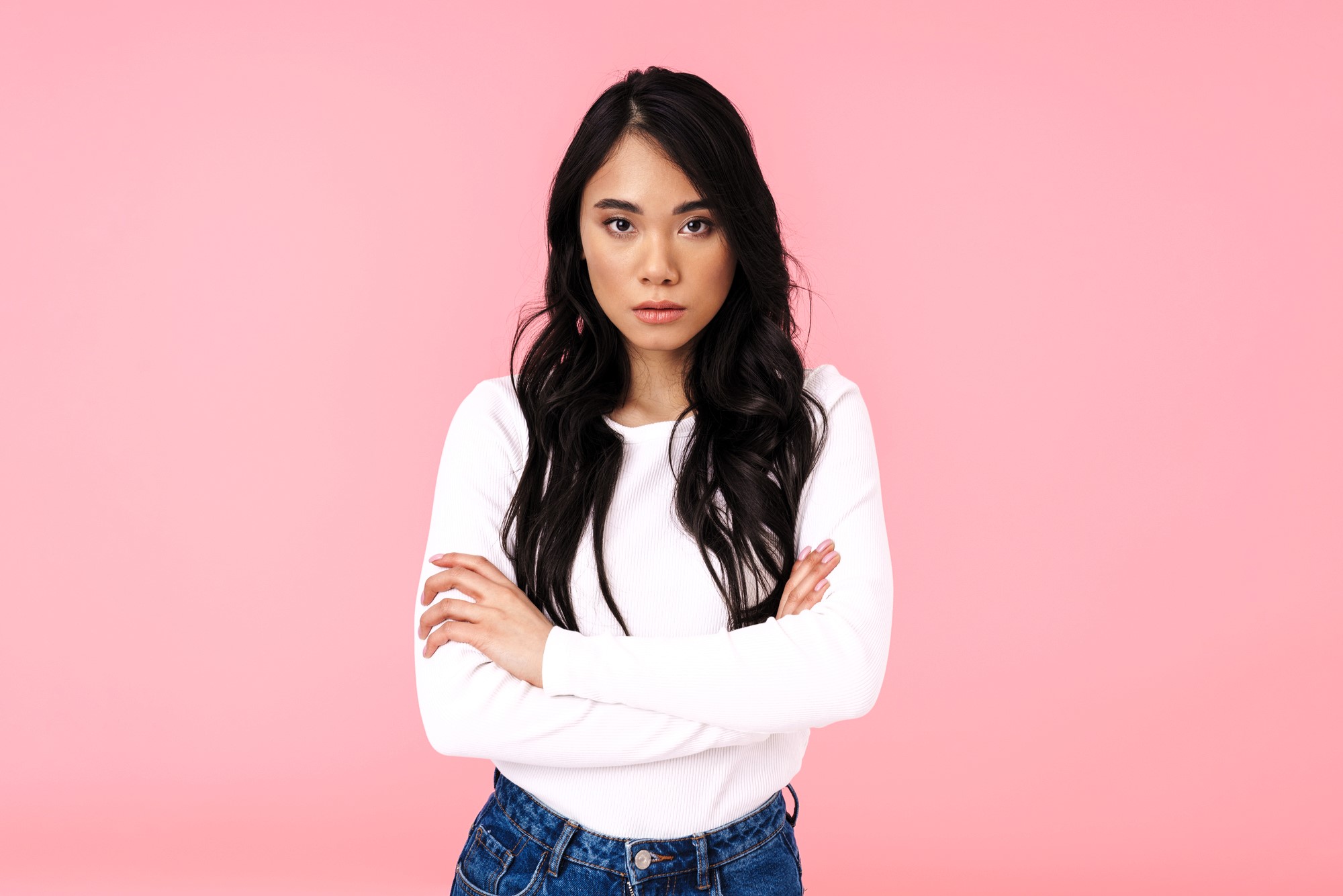 A woman with long dark hair stands against a pink background, wearing a white sweater and blue jeans. She has her arms crossed and a serious expression.