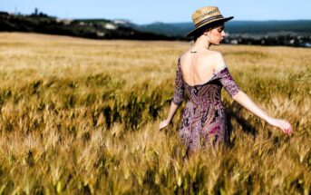 A woman in a patterned dress and straw hat walks through a golden wheat field. Her back is turned towards the camera, and the sky is clear. She gently touches the wheat stalks with her hand.